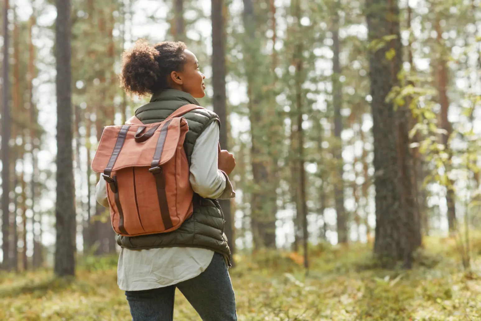 Back view portrait of young African-American woman with backpack enjoying hiking in forest lit by sunlight.