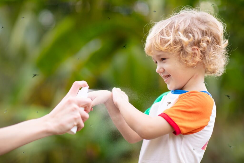 A child receives mosquito spray on their arm as mosquitoes swarm around.